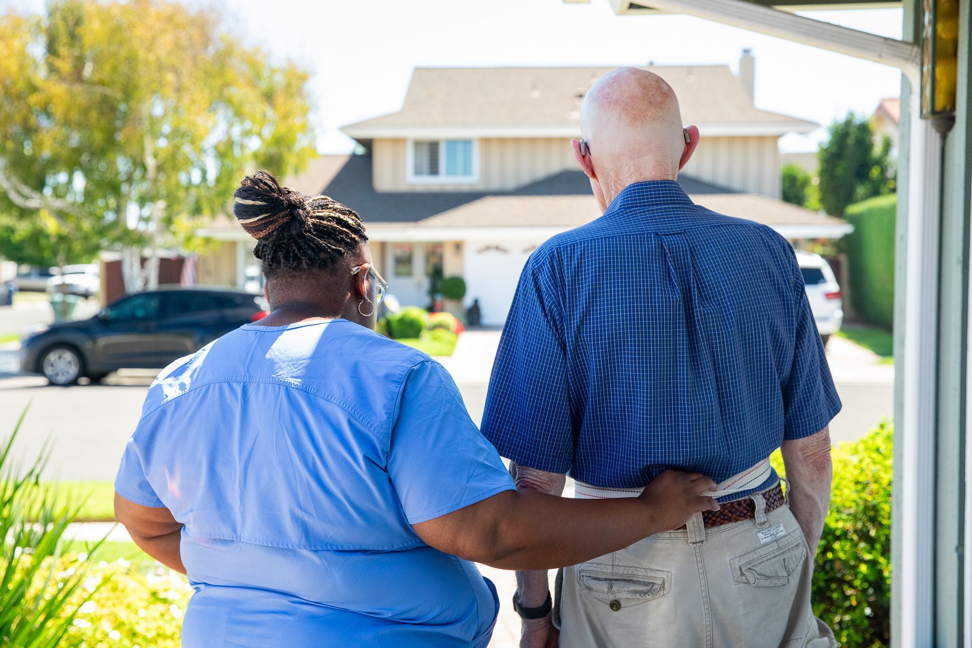 Two people standing on a porch, one supporting the other, with a suburban street and house in the background