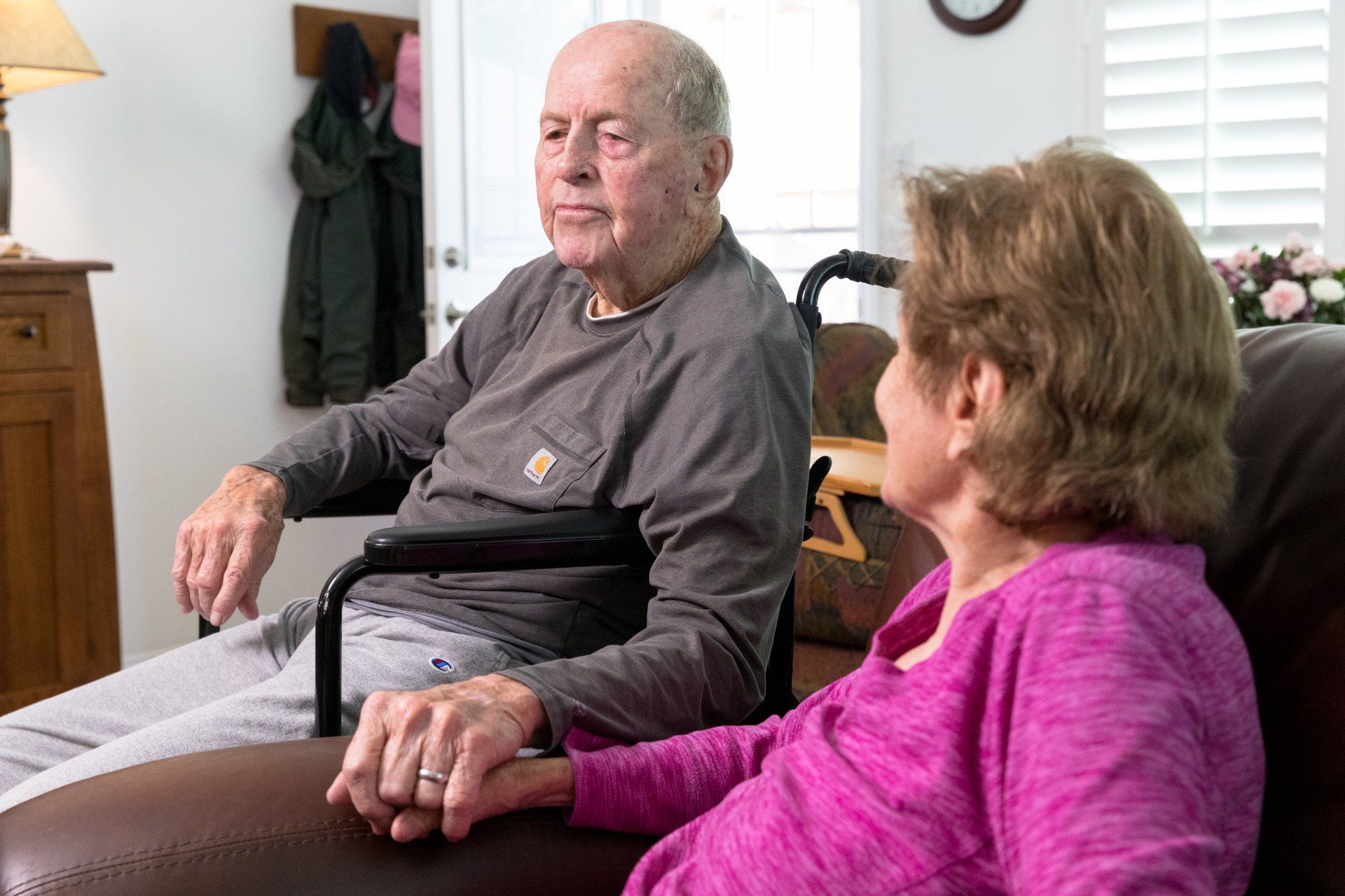 Older couple sitting indoors, man in wheelchair, holding hands. Brown leather couch, casual clothes.