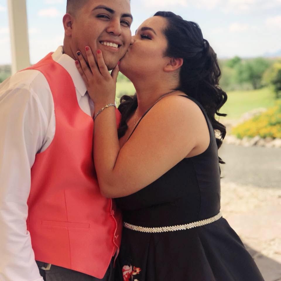 Two people smiling outdoors, one kissing the other's cheek on a bright terrace.