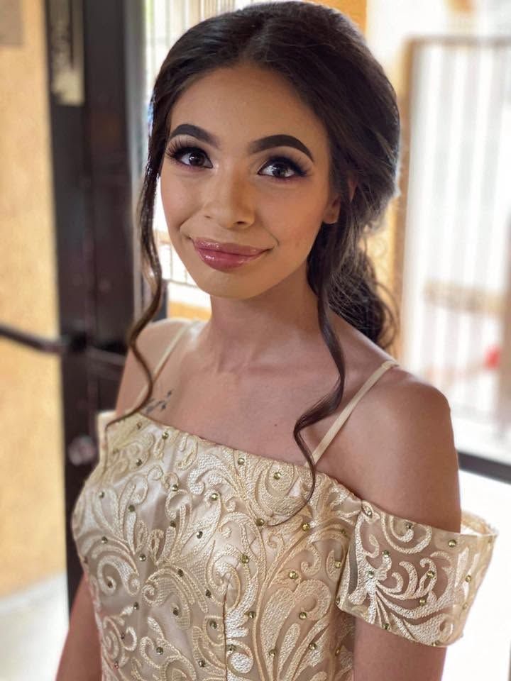 Woman in a gold off-the-shoulder dress, smiling indoors near a window.