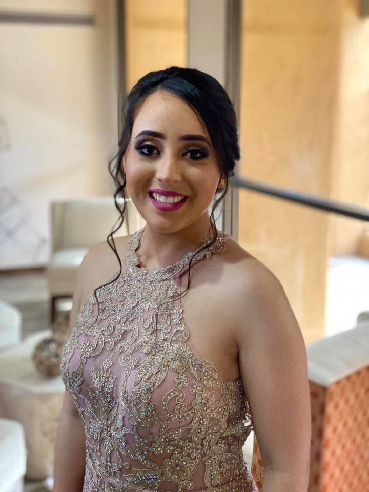 Smiling woman in a sparkling pink gown, standing indoors near a staircase and curtains.