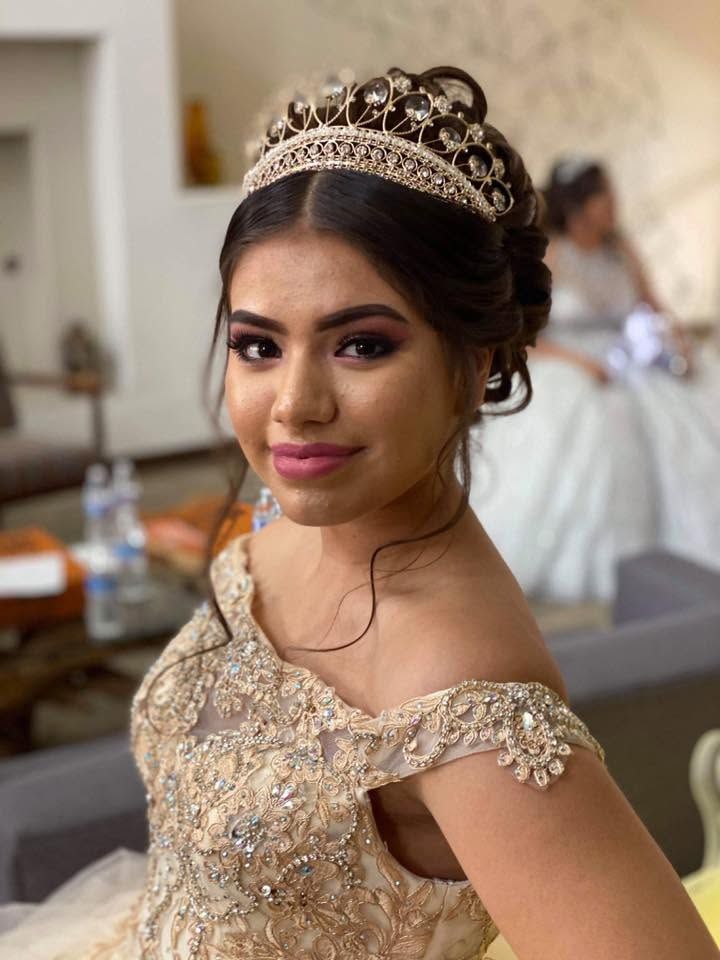Woman in a gold gown and tiara, smiling in a softly lit indoor setting.