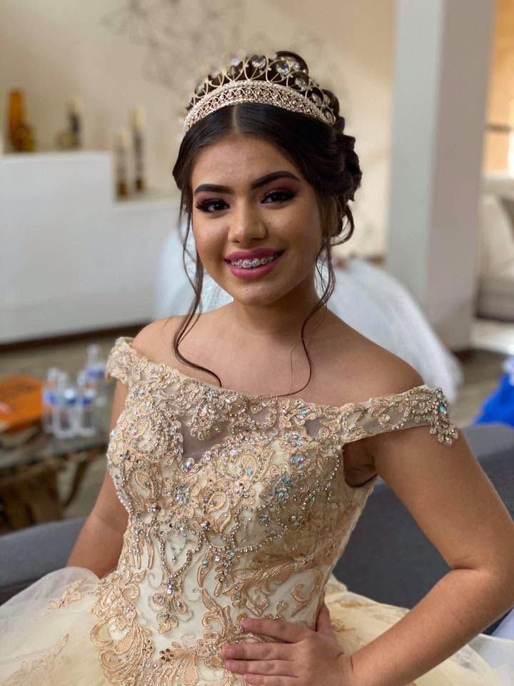 Smiling woman in a gold off-shoulder gown and tiara, standing indoors.