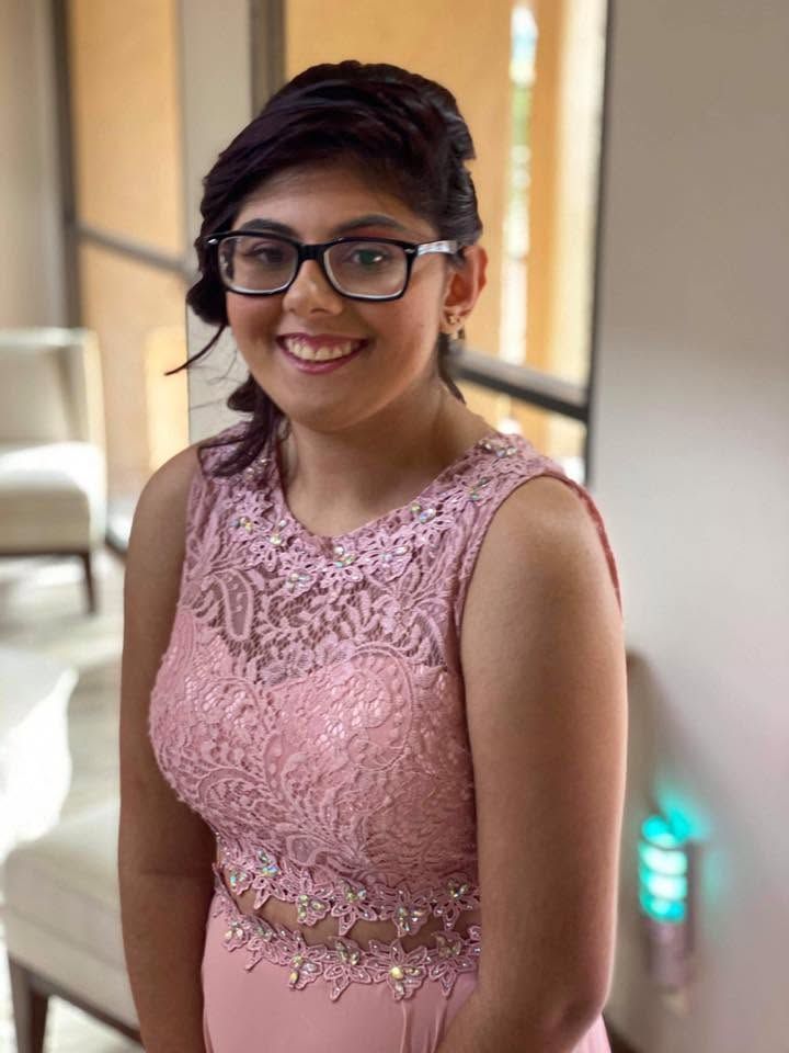 Woman in a pink lace dress smiling indoors, standing near a window.