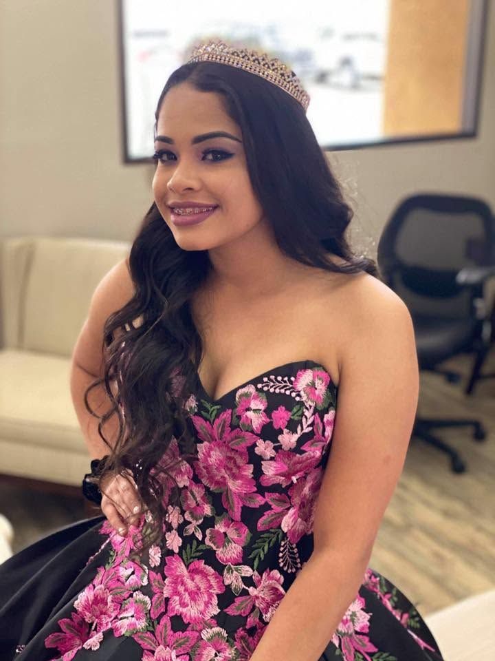Woman in a pink floral gown and tiara, smiling indoors against a blurred office background
