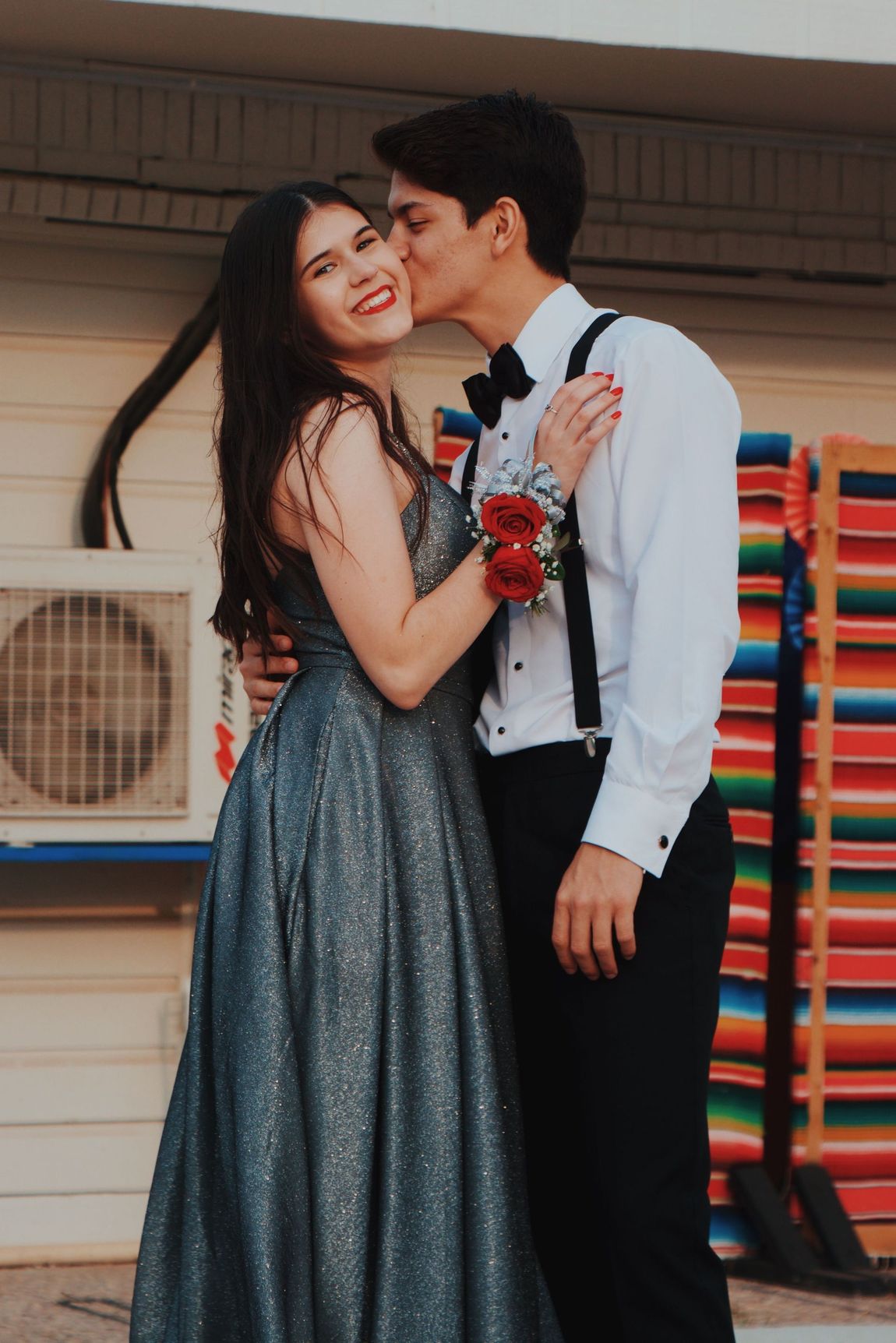 Couple in formal attire embracing outdoors, with the man kissing the woman’s cheek.