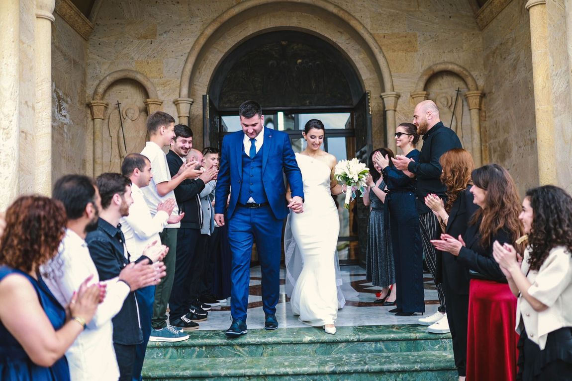 Bride and groom exit a stone church, surrounded by applauding guests.