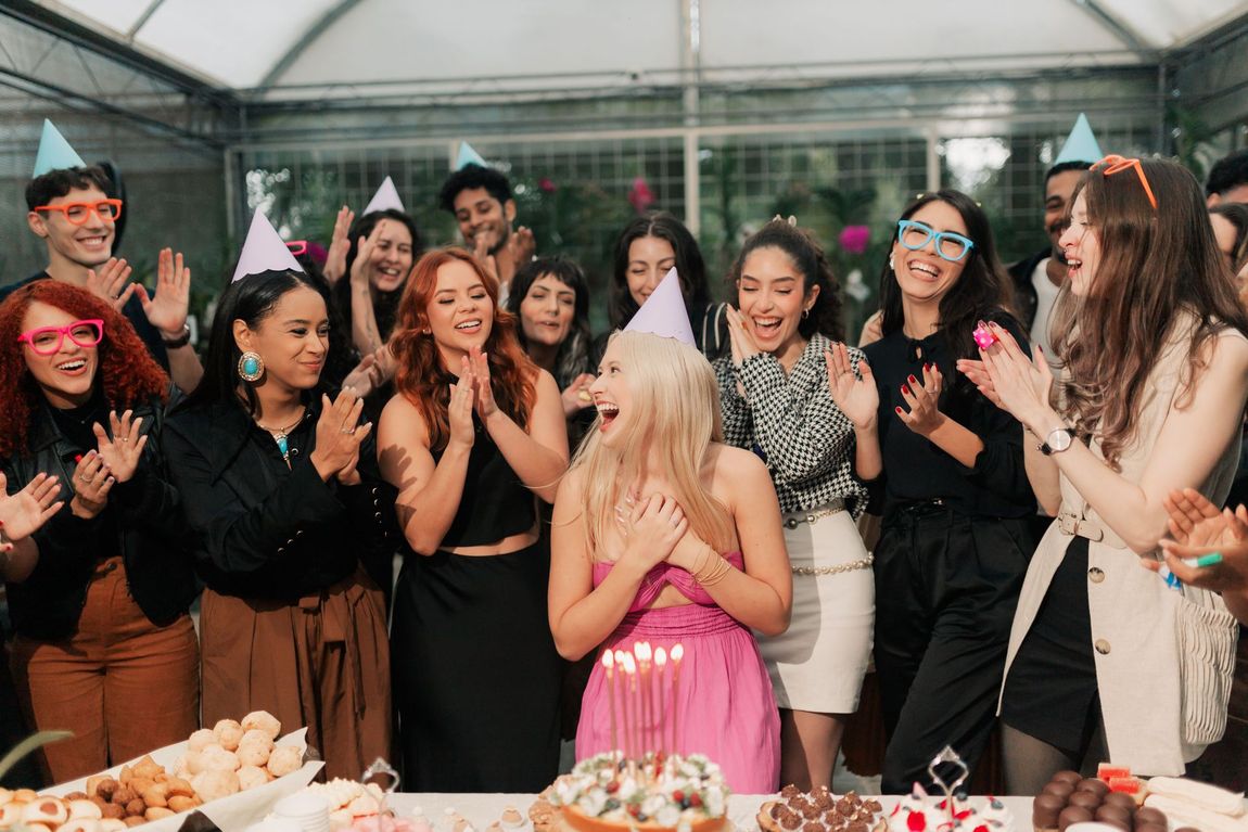 People at a party wearing hats and dresses, cheering around a table with cake and food.