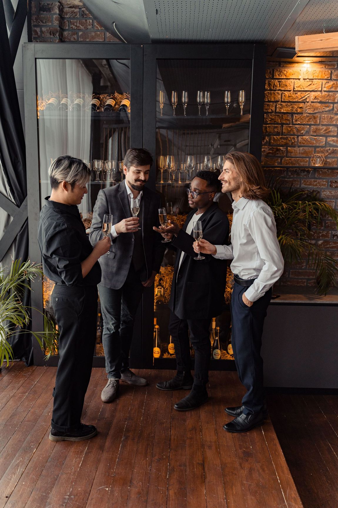 Four people chatting with drinks on a wooden patio outside a brick building at night.