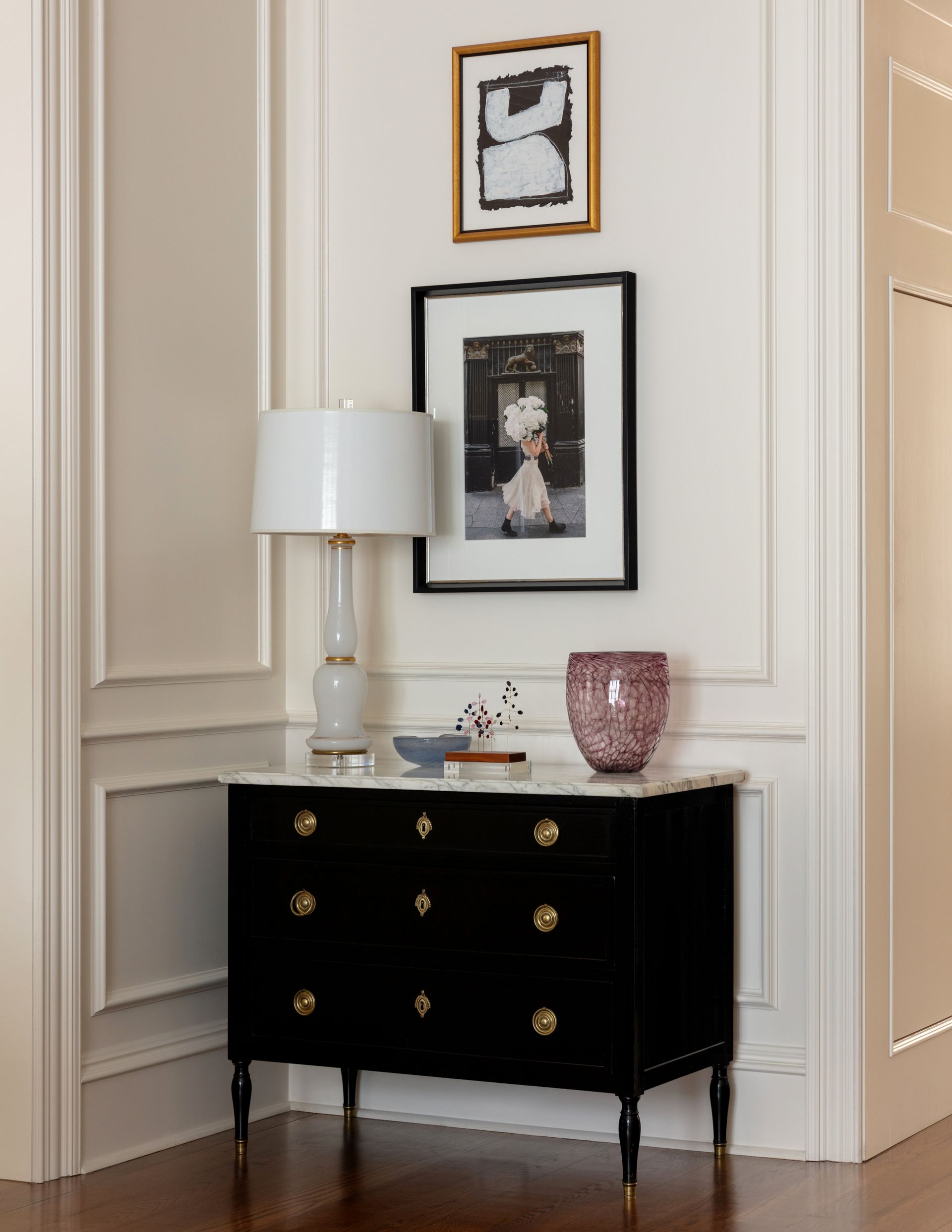 Black dresser with gold knobs, white lamp, and framed art in a paneled hallway corner.