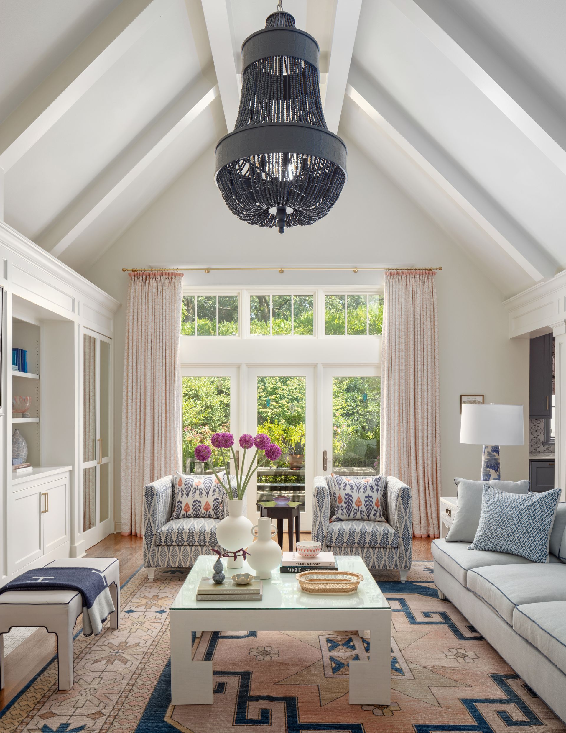 Bright living room with vaulted ceiling, black chandelier, large window, blue-and-white furniture, and floral accents