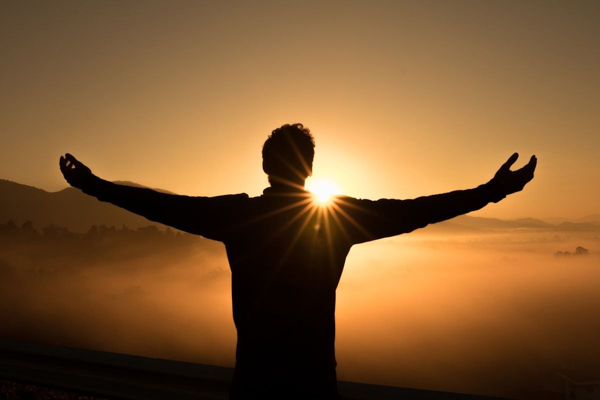 Silhouetted person with arms outstretched at sunrise, standing before a glowing orange sky