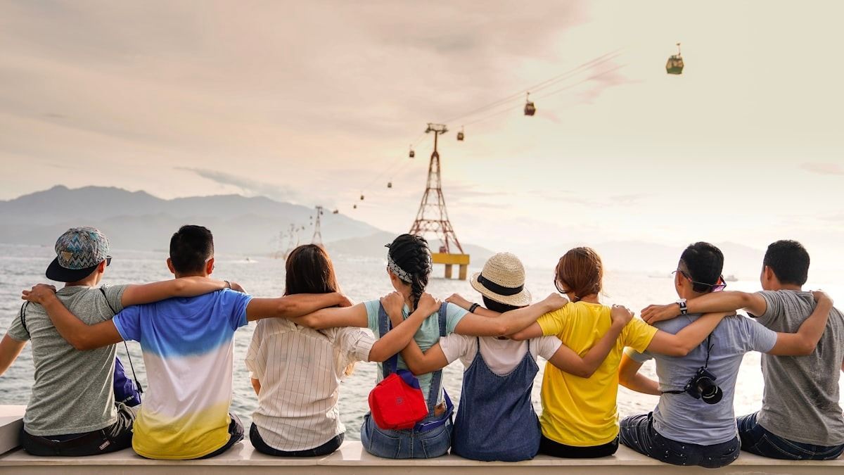 Group of friends sitting arm in arm, facing a cable car over a hazy mountain landscape at sunset