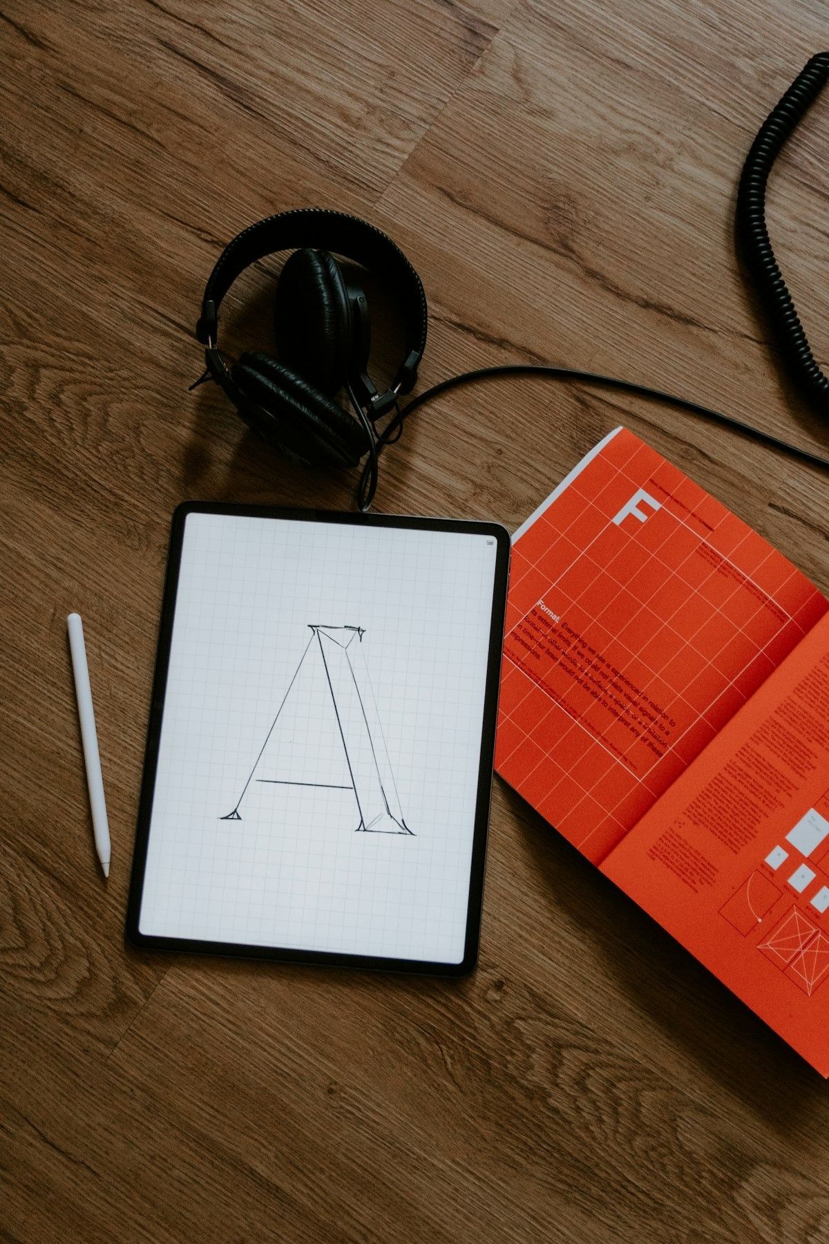 Flat-lay of black headphones, a notebook with a sketched letter A, and an orange notebook on a wooden desk