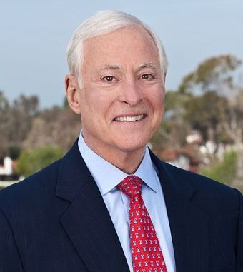 Portrait of a smiling man in a navy suit and red tie outdoors, with trees and sky in the background