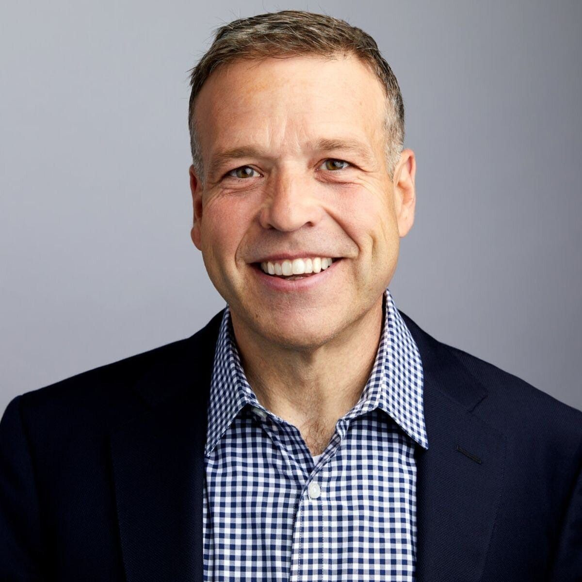 Smiling man in a navy blazer and blue checkered shirt against a gray background