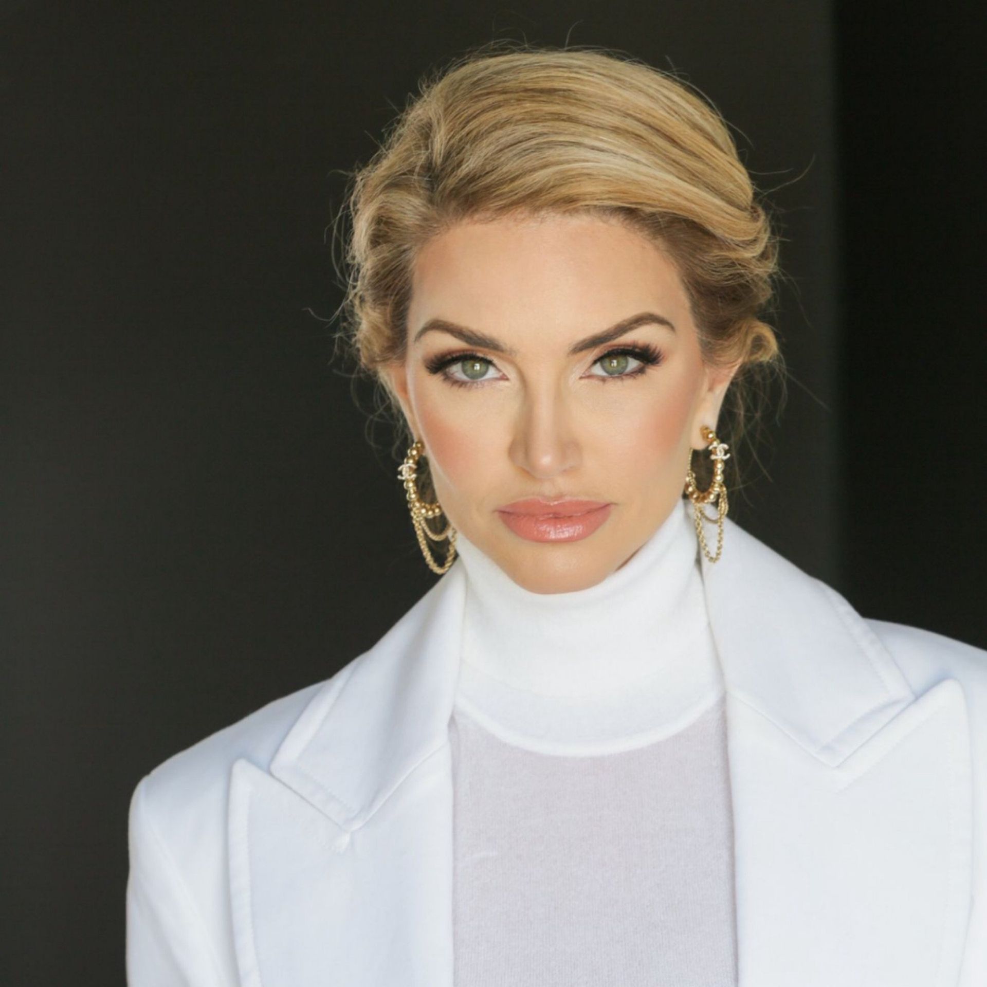 Portrait of a woman in a white blazer and turtleneck against a dark background, wearing gold earrings