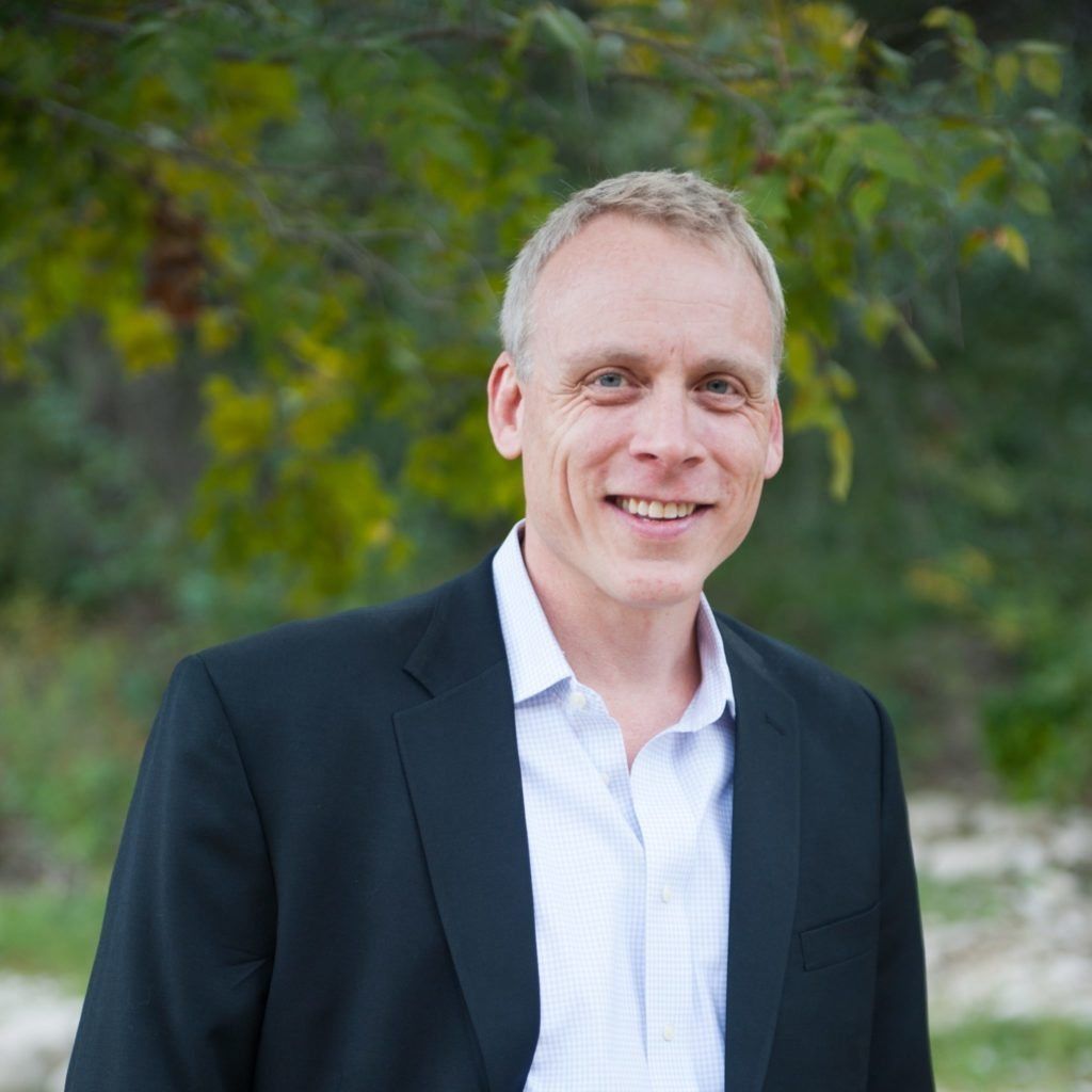 Smiling man in a black blazer and white shirt standing outdoors with green foliage behind him