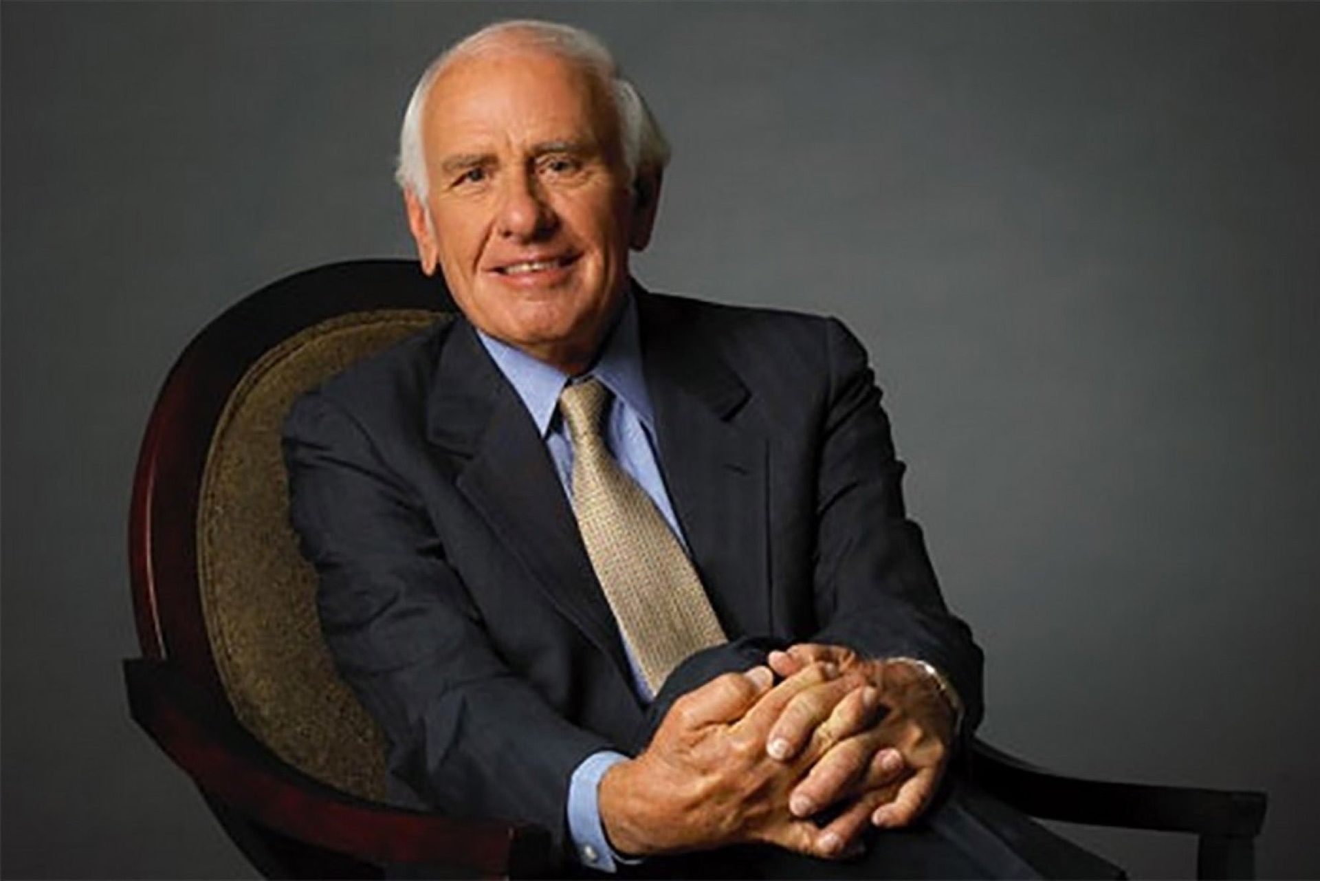 Smiling man in a dark suit seated in a chair against a gray background