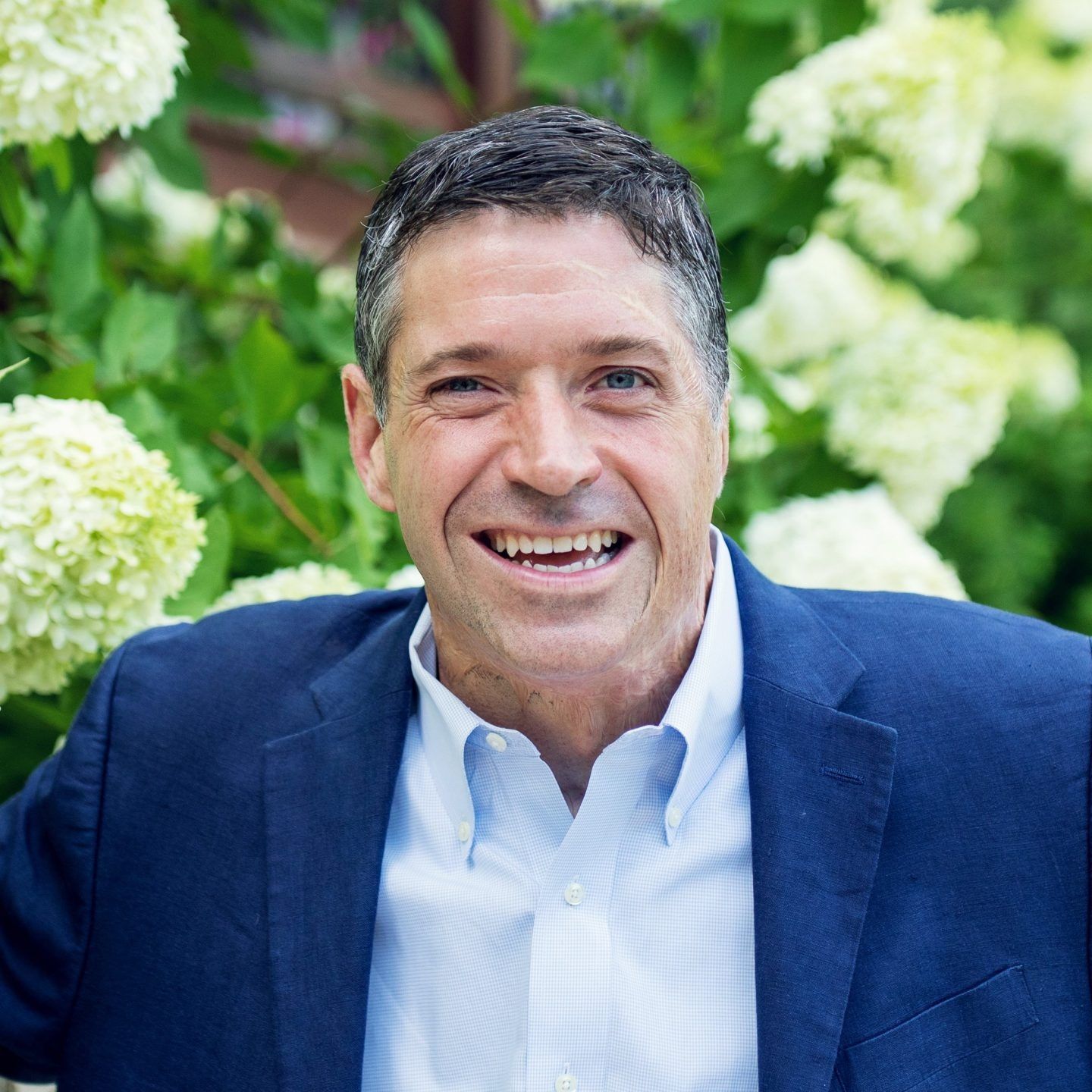 Smiling man in a blue blazer and white shirt outdoors in front of green flowers
