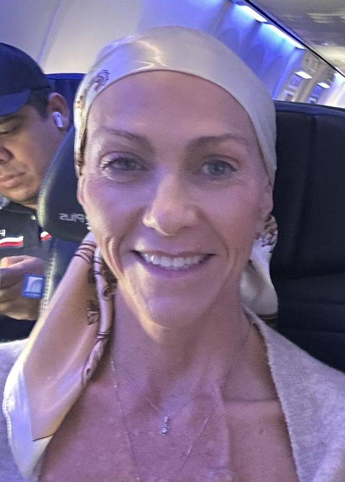 Smiling person in a vehicle seat, wearing a light head covering and beige top, with airplane cabin lighting.