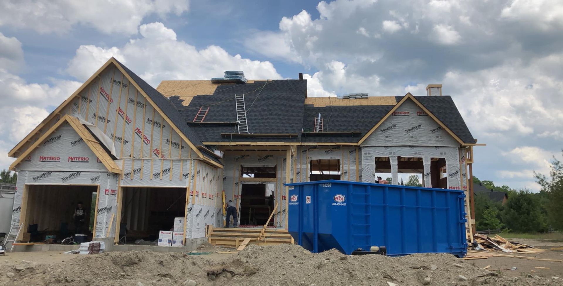 Construction d'une maison neuve avec un conteneur à déchets bleu dans la cour avant ; toiture et revêtement partiellement terminés.