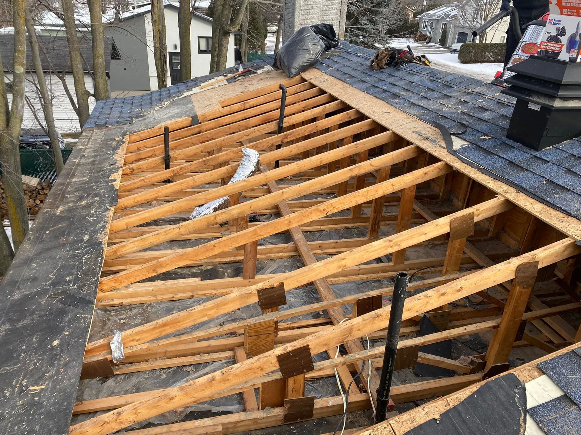 Travaux de toiture en cours, chevrons et revêtement apparents, bardeaux bleus, cheminée visible.