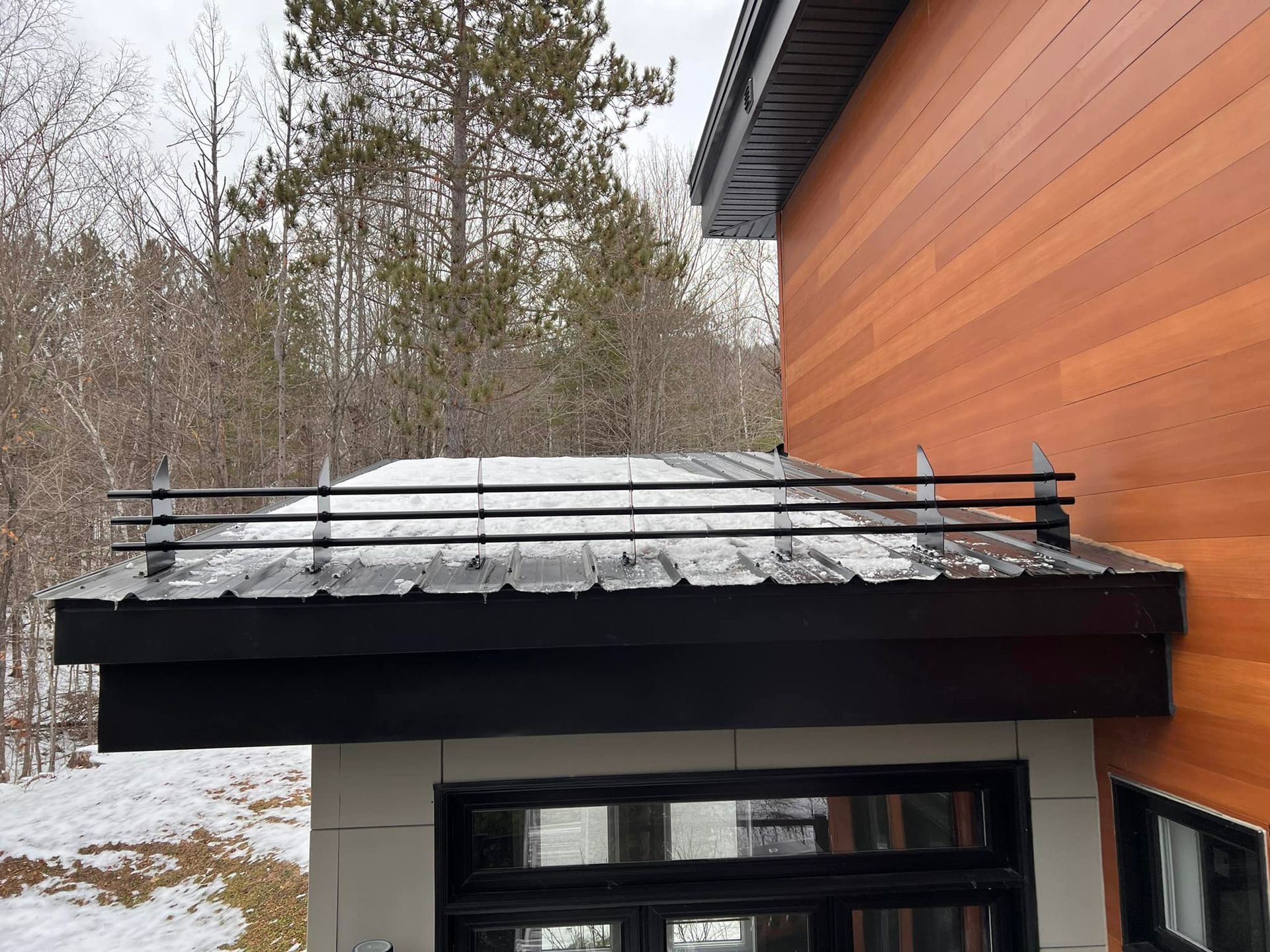 Toit noir avec rambarde, recouvert de neige, au-dessus des fenêtres d'un bâtiment moderne à bardage en bois, avec la forêt en arrière-plan.