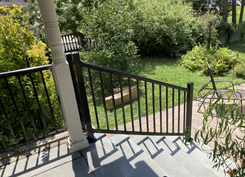 Black railing on porch steps, leading to a grassy yard with trees.