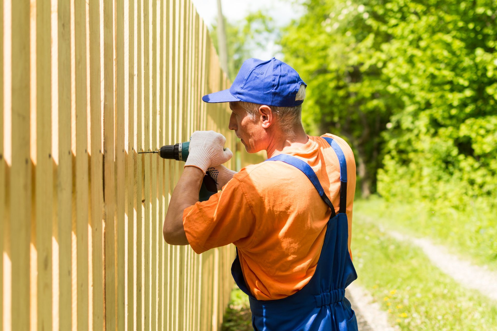 Man in overalls and cap using a drill to install a wooden fence.