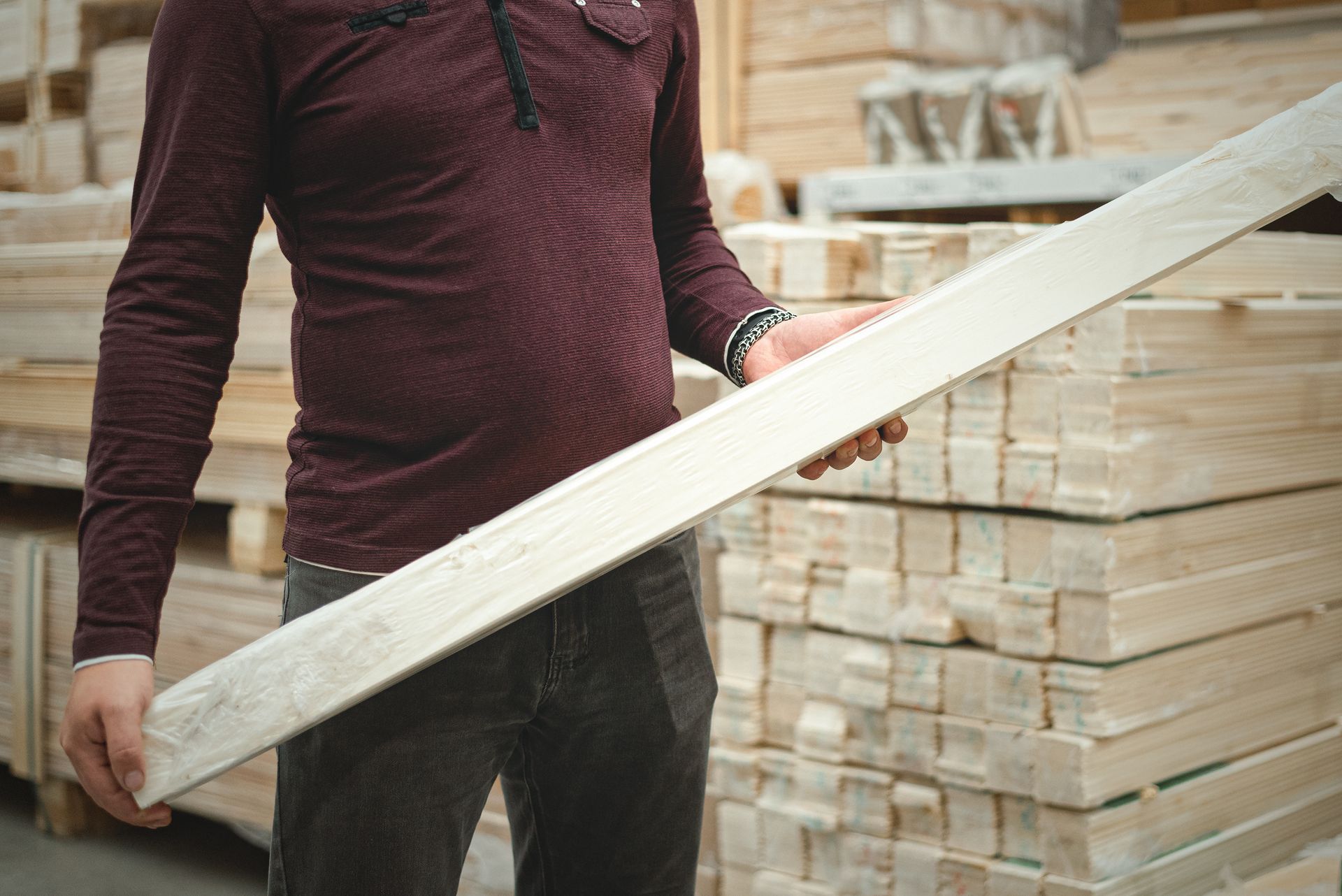 Man in burgundy shirt holding a wooden plank in a lumberyard.