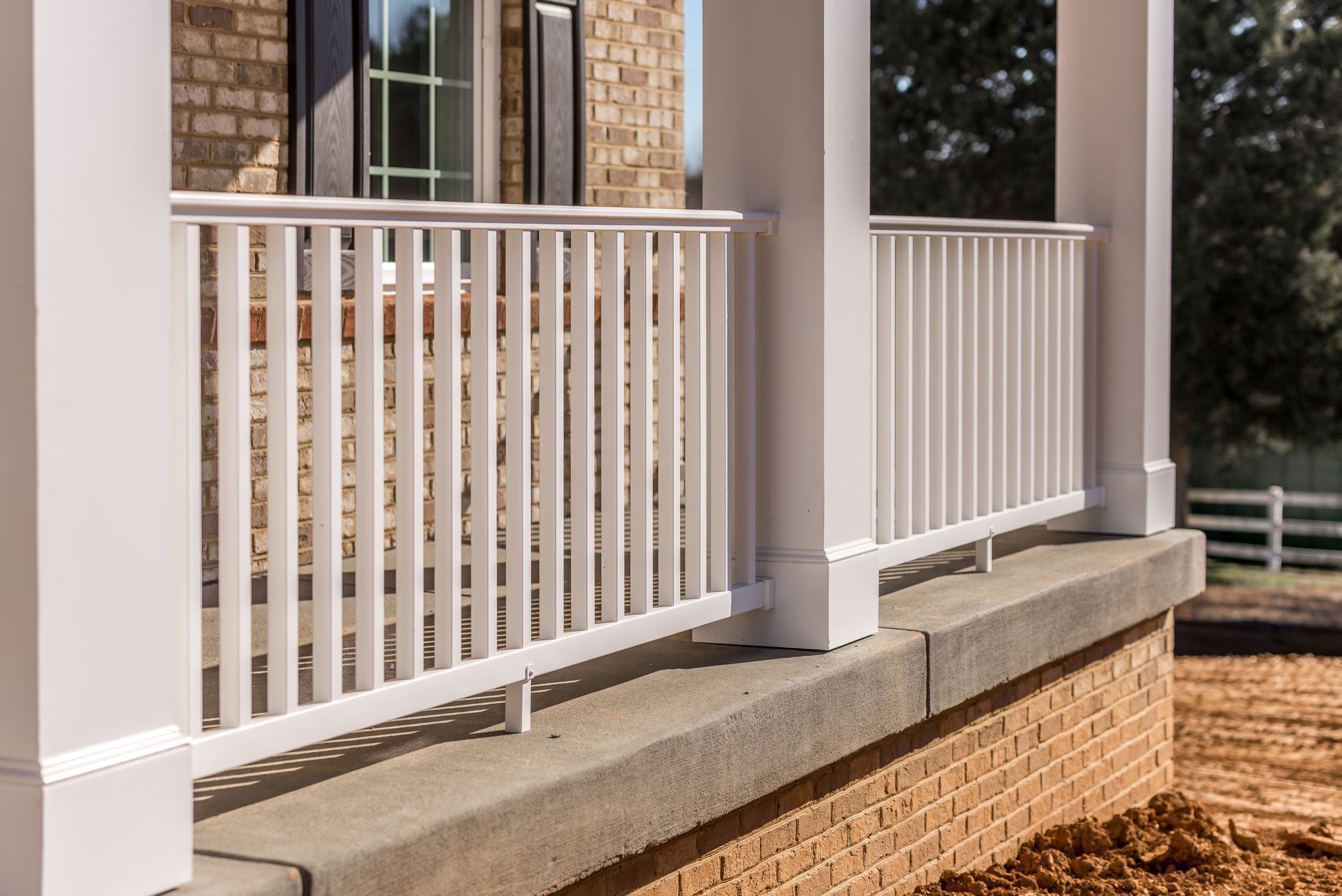White picket fence in front of a brick house. Fence surrounds brown mulch in a neighborhood.