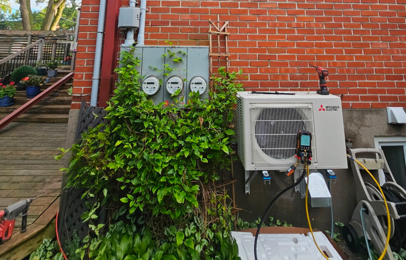 Unité de climatisation extérieure à côté d'un mur de briques, avec des compteurs électriques et un buisson feuillu devant une terrasse en bois.