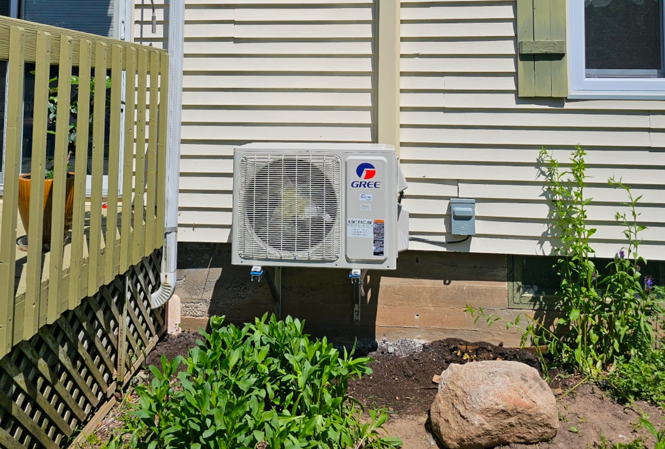 Unité de climatisation extérieure à côté d'une maison, avec des arbustes et une rambarde de terrasse en bois à proximité.