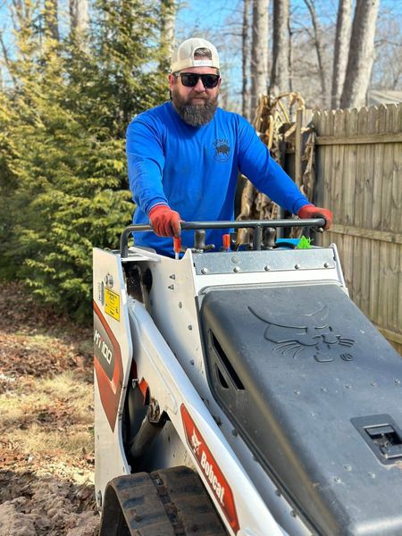 Worker in blue jacket and red gloves operating a skid steer on a dirt path beside a fence.