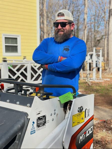 Bearded man in blue hoodie and sunglasses standing with arms crossed beside a snow blower outdoors