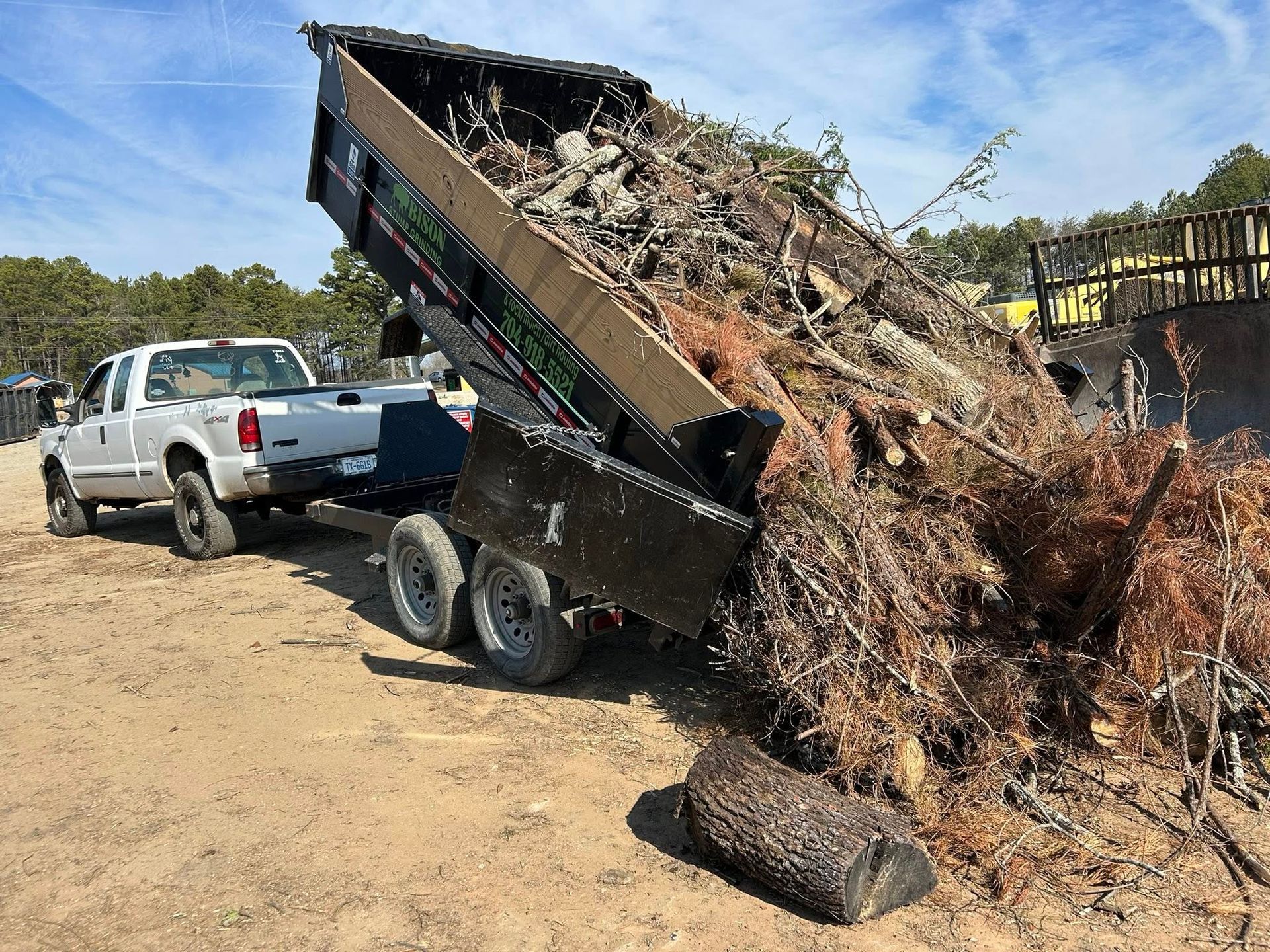 Dump trailer tilting with a large pile of yard debris beside a white pickup on a dirt lot under a blue sky