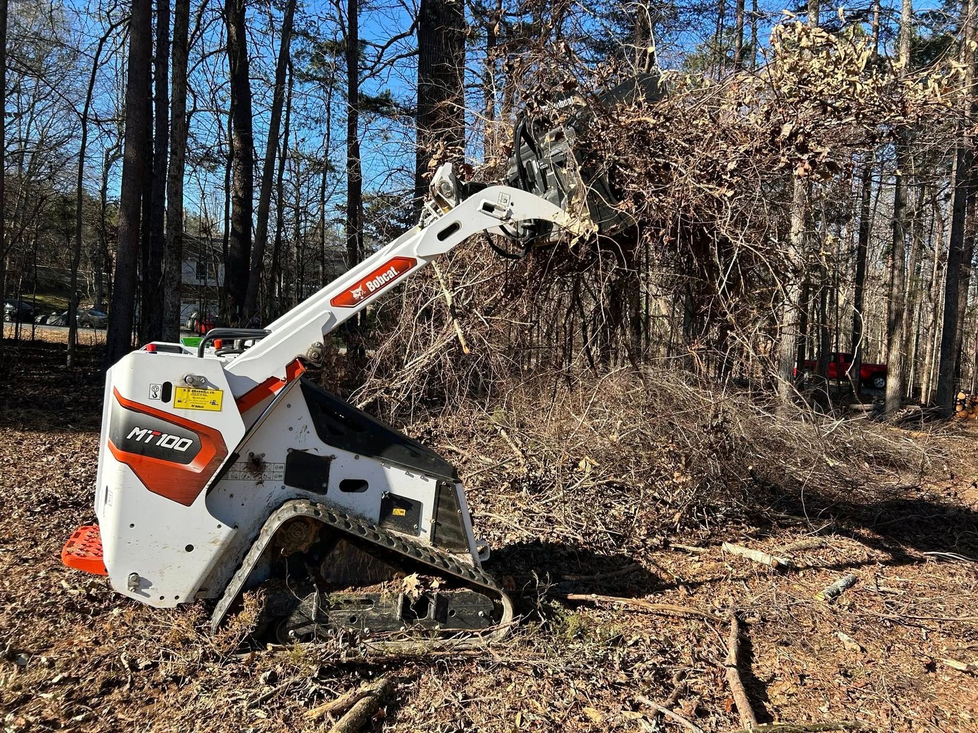 White Bobcat compact loader clearing brush in a wooded area under a blue sky