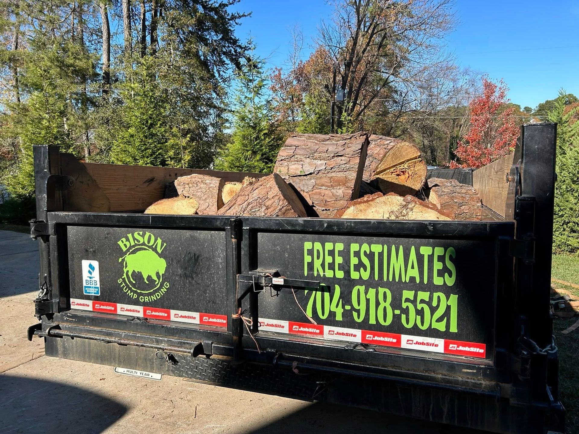 Black dumpster trailer filled with yard debris, with “Free Estimates” sign and phone number in a sunny wooded area