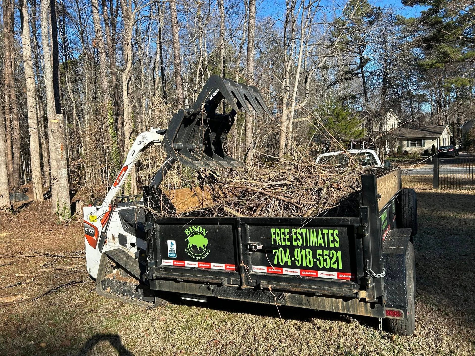 Black dump trailer filled with yard debris and a small excavator in a wooded driveway
