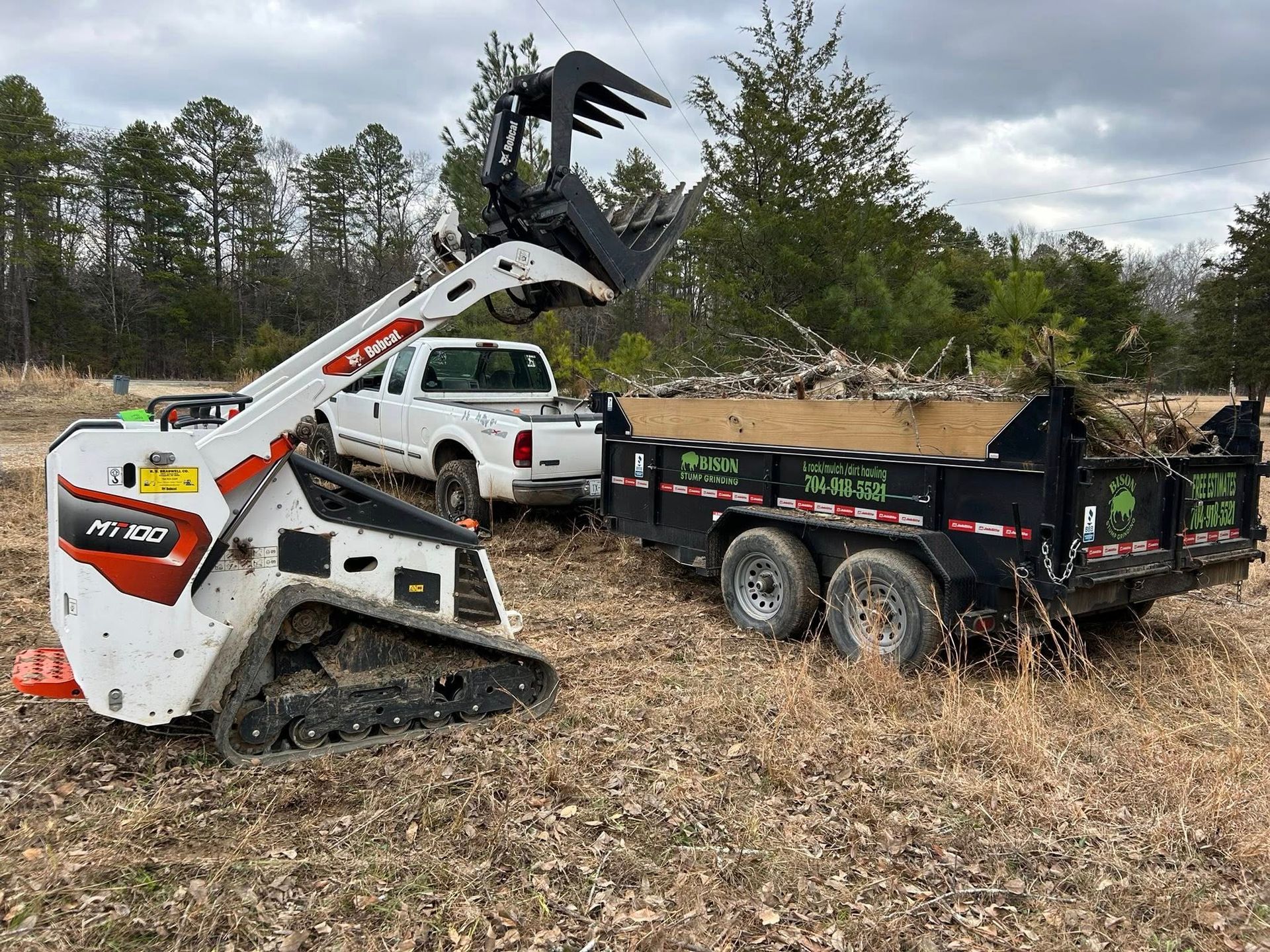 White skid-steer loader lifting debris into a black dump trailer in a wooded field