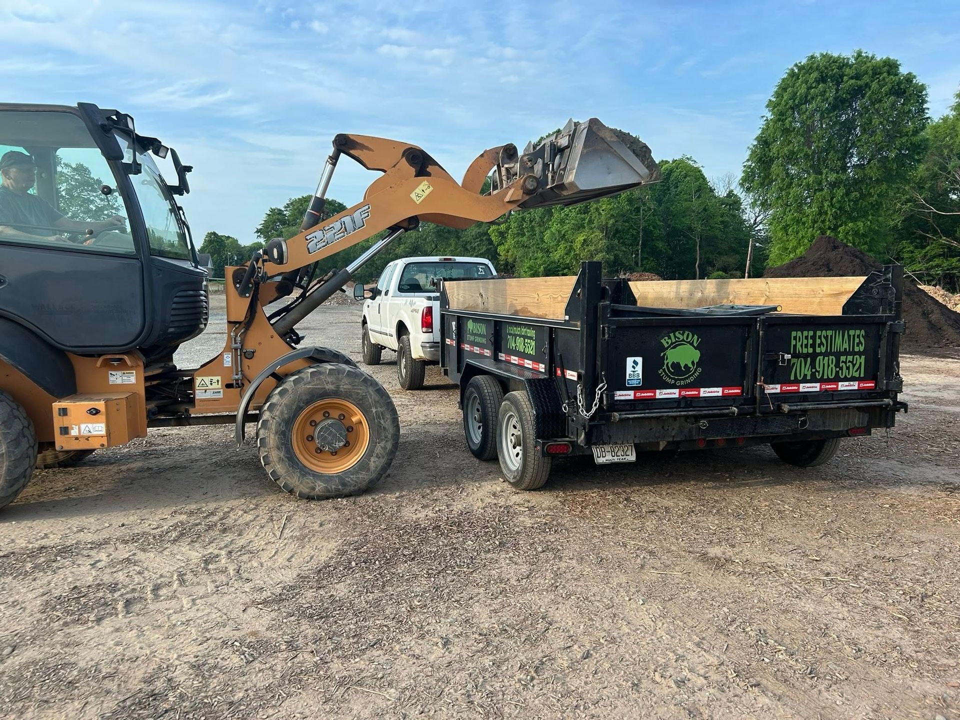 Loader dumping dirt into a black dump trailer on a gravel site, with other construction equipment nearby.