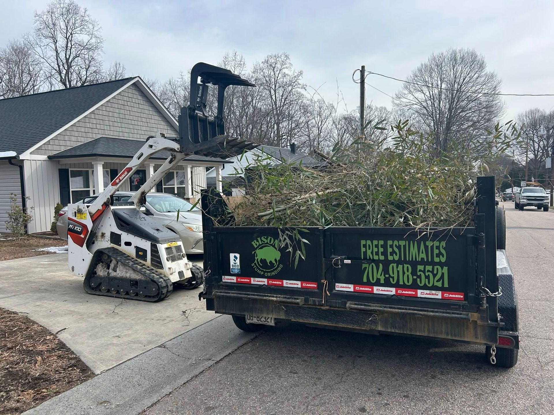 Work truck with trailer and skid steer at a house driveway during tree removal cleanup