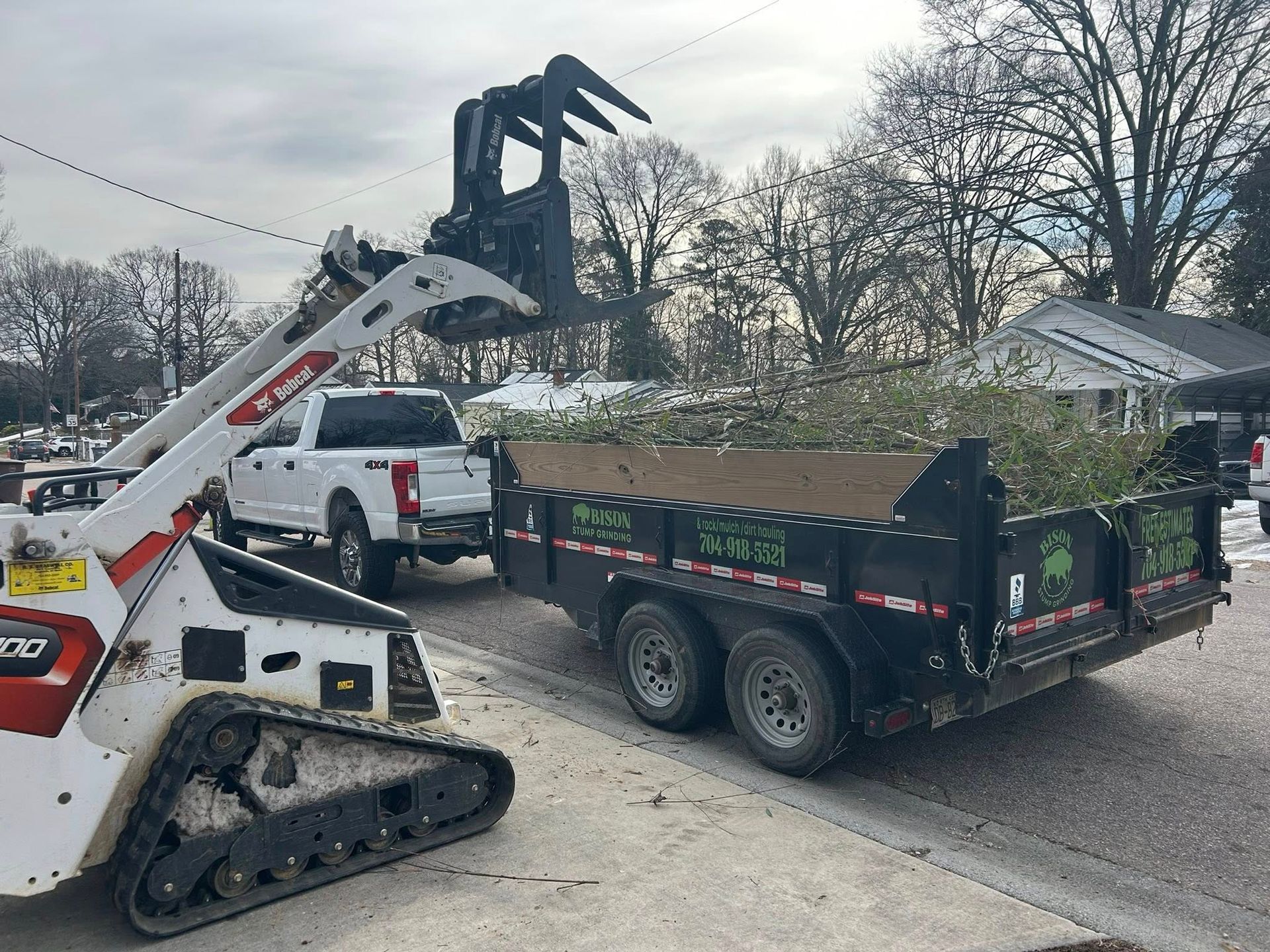 Construction loader lifting debris into a black dump trailer on a street, with a white truck nearby