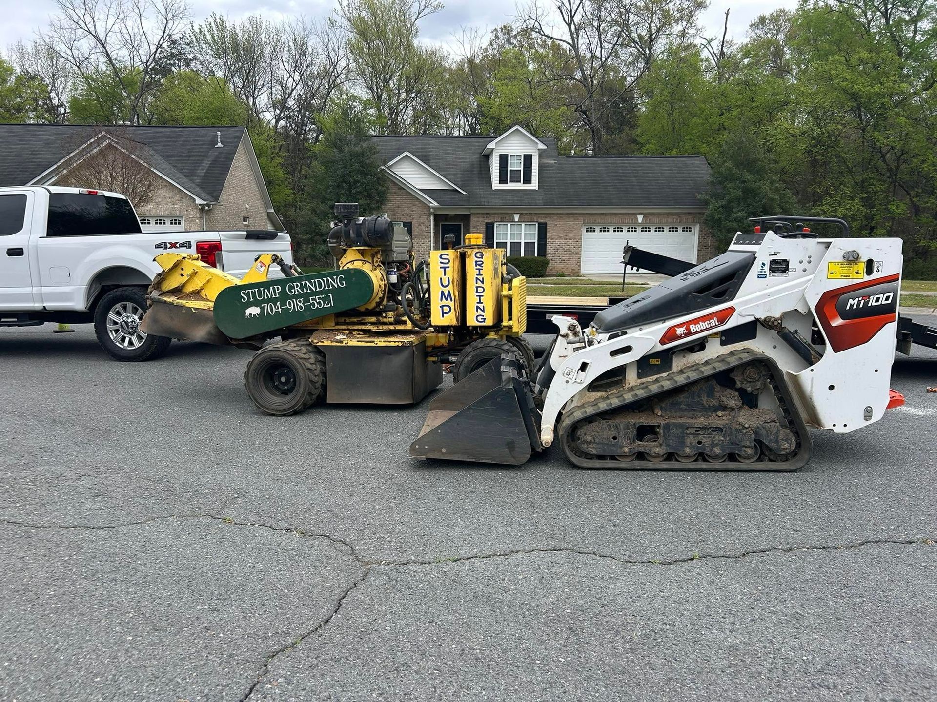 Two compact construction machines parked on a street in front of houses and trees.