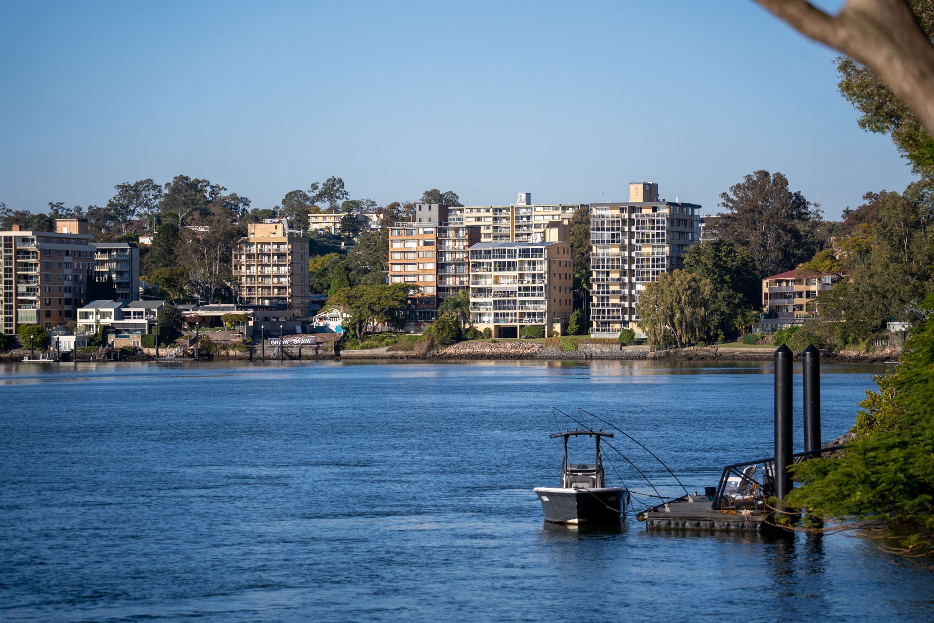 Riverside city skyline across blue water with docked boat and trees in the foreground