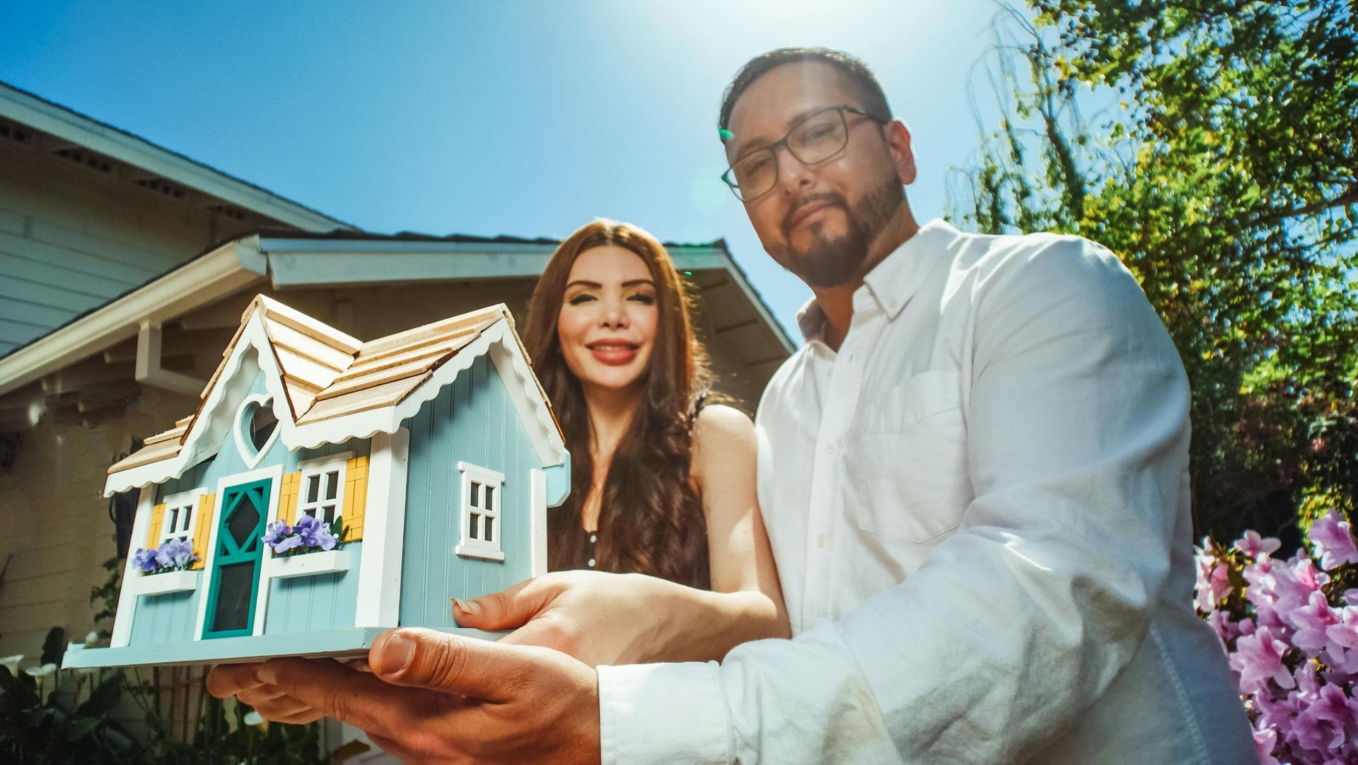 Man in white shirt and woman stand before a small pastel house under a sunny blue sky.