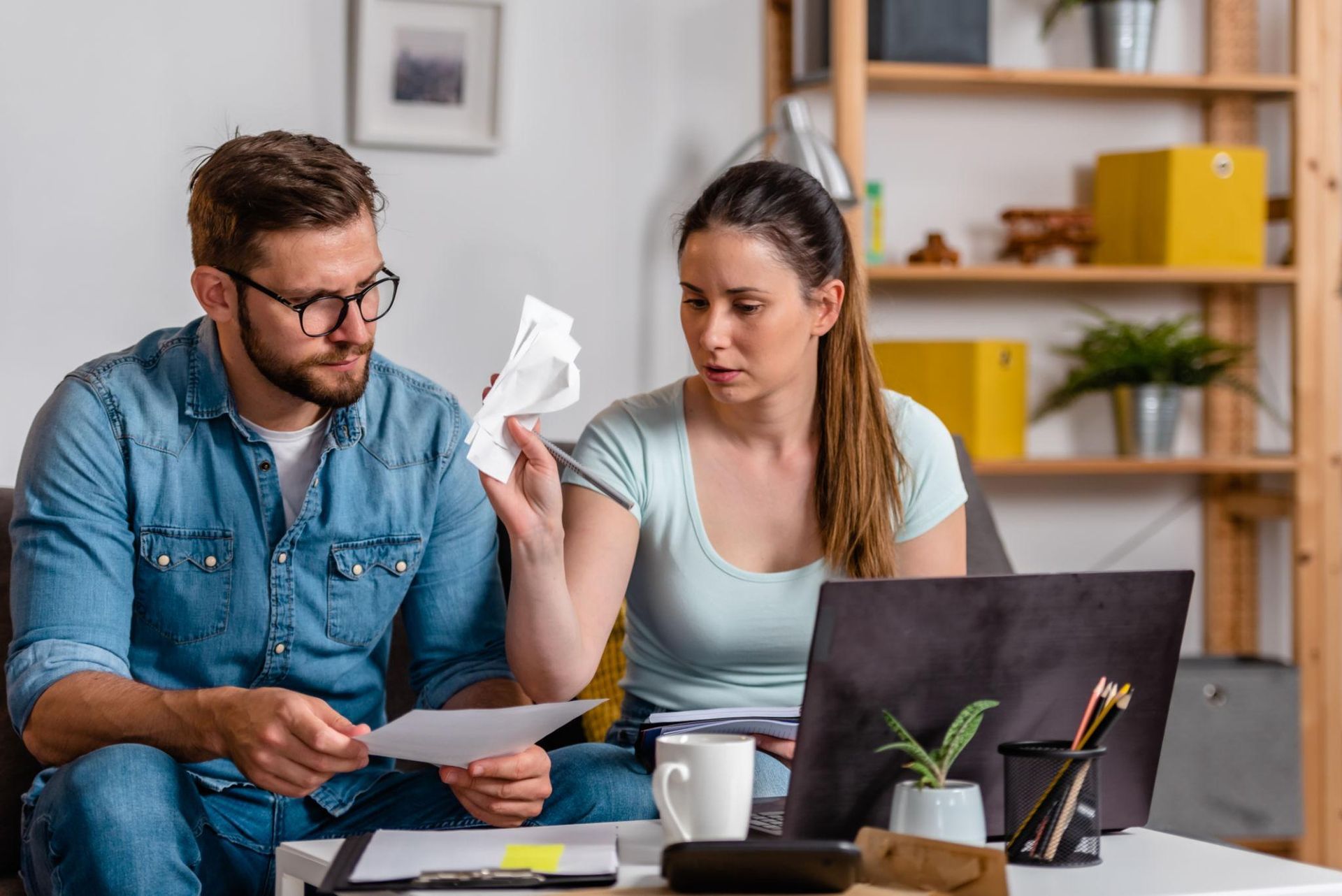 Two people review bills at a table with a laptop, looking concerned and holding paperwork.
