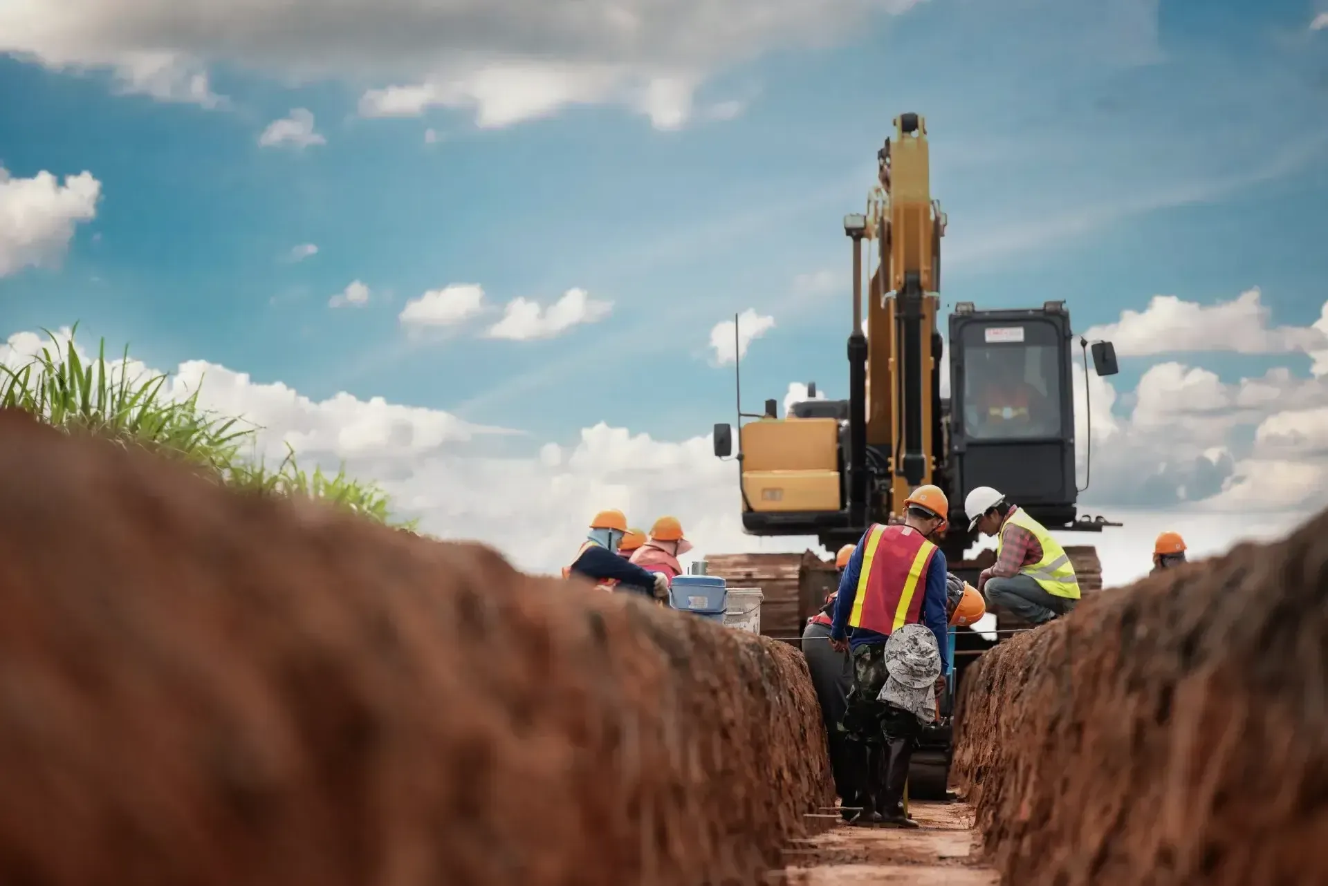 Ouvriers travaillant dans une tranchée de terre avec une excavatrice jaune en arrière-plan.