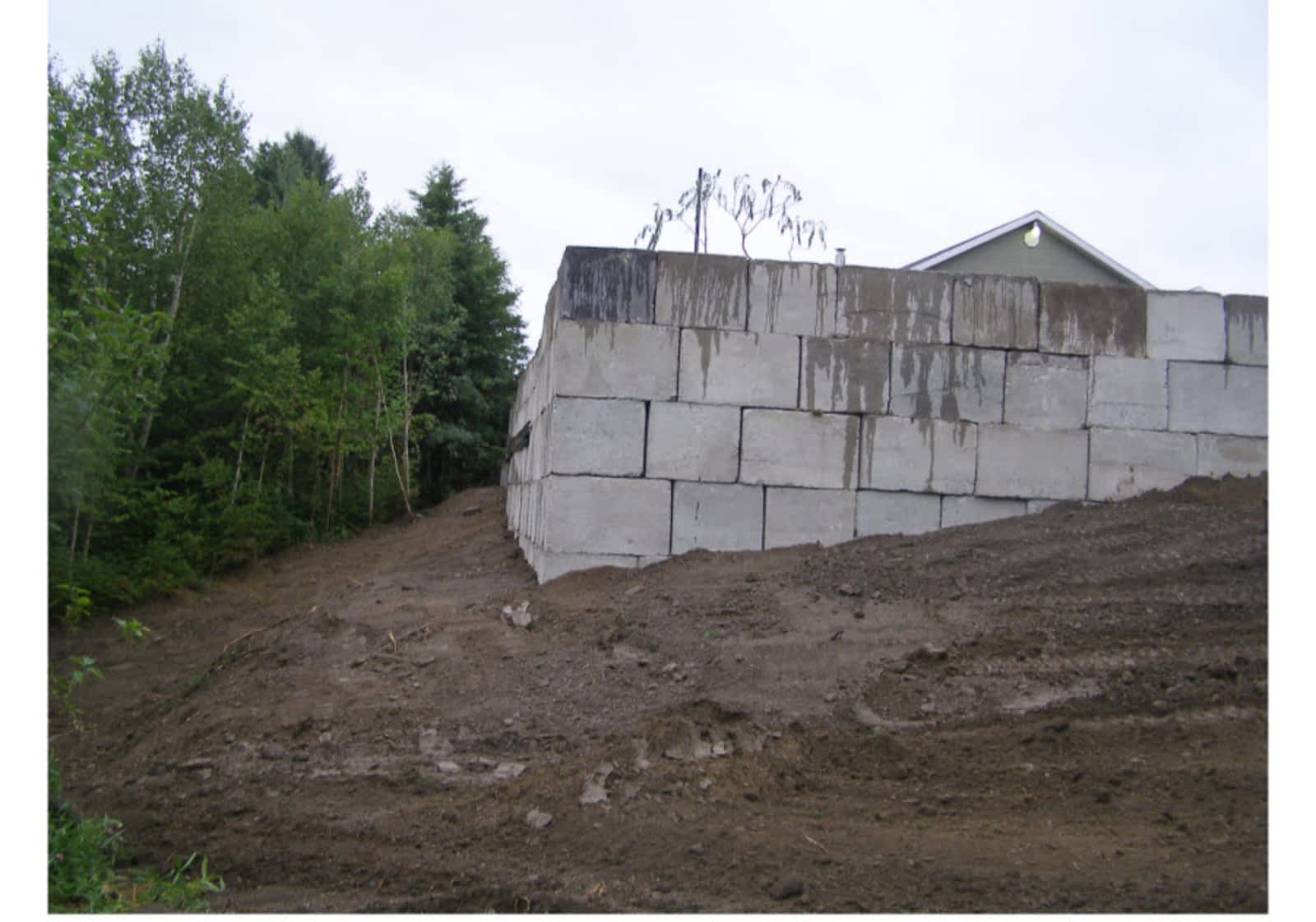 Grand mur de soutènement en blocs de béton sur une pente de terre nivelée près d'une forêt.